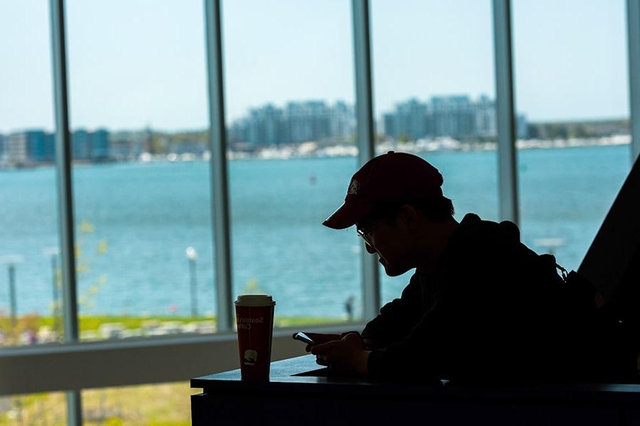 Silhouette of student in baseball hat looks at cellphone next campus center window.
